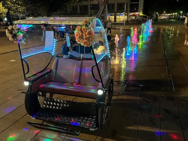 Rikscha vor Regenbogen-Brunnen bei Nacht.
